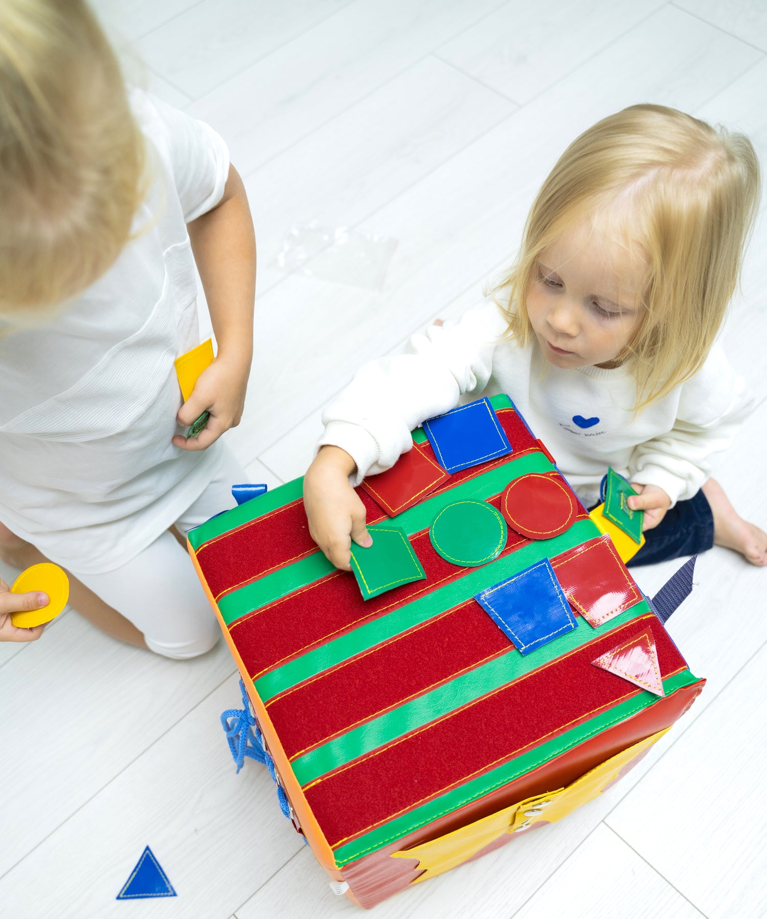 Two young children playing with a large colourful felt quiet book/busy cube, shape sorter soft toy for toddlers' fine motor skills, Montessori educational play cube for kids' development.