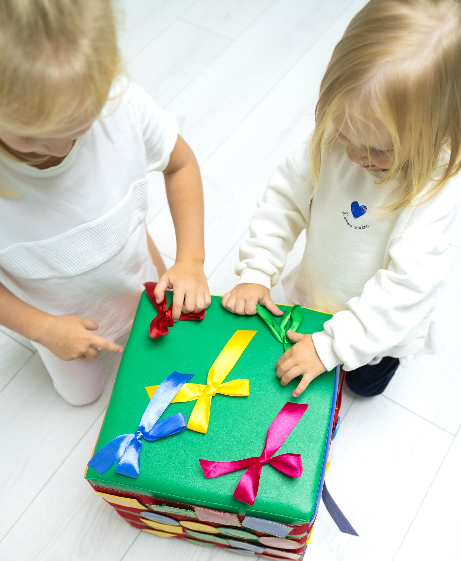 Two toddlers playing with a green busy cube or quiet book soft toy, Montessori educational toy for fine motor skills and lacing practice, large fabric activity cube for children's development.
