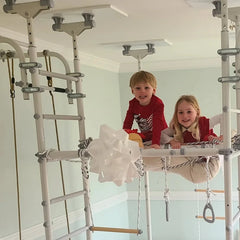 Two children playing on a metal play structure in a room with a white wall and green curtain.