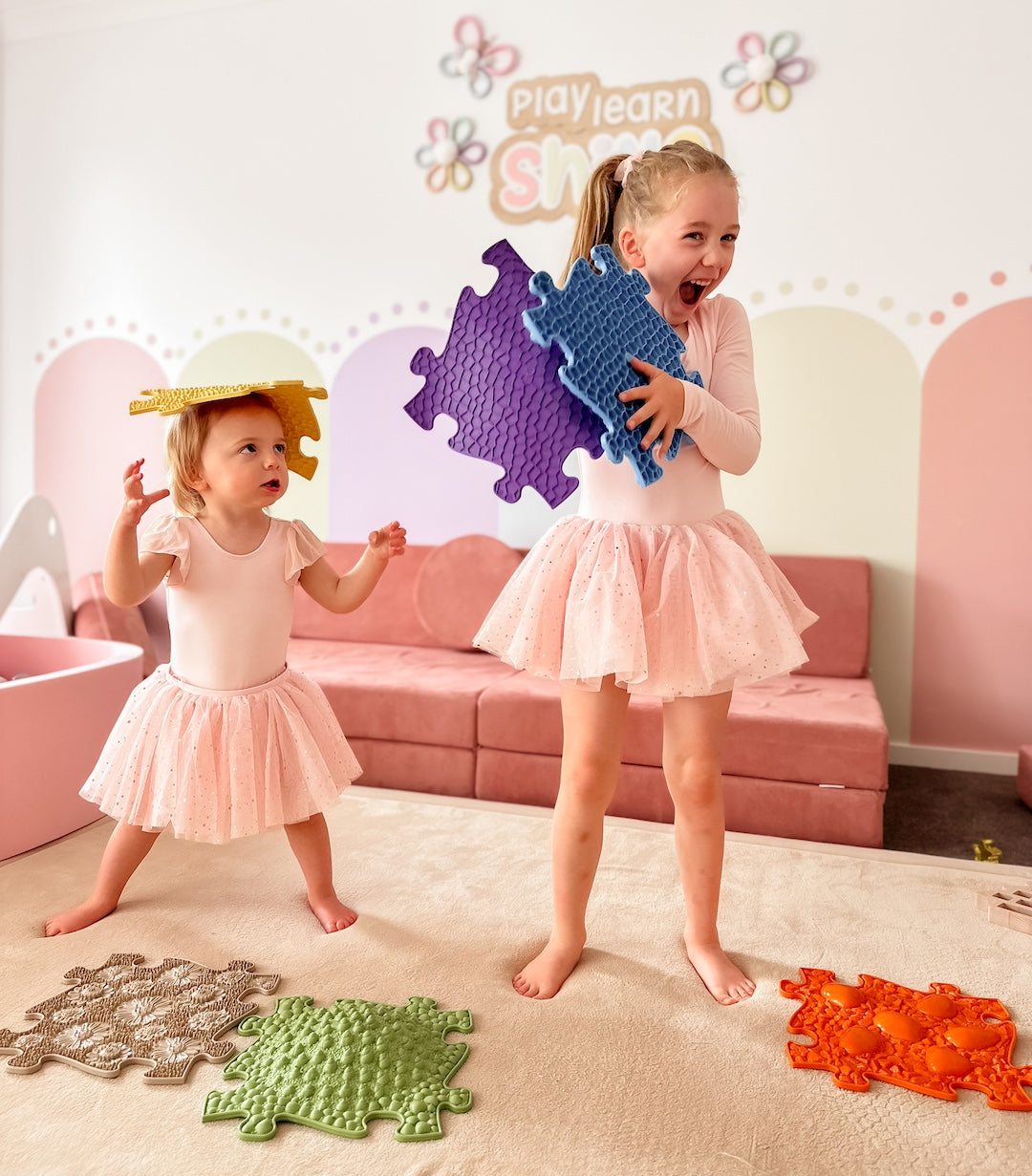 Two young girls, wearing pink dresses, play with colorful foam puzzle pieces from Muffik. One girl balances a piece on her head; the other holds one and laughs. The background shows a wall with the words "play, learn, share." These sensory activities bring joy to their endless imagination.